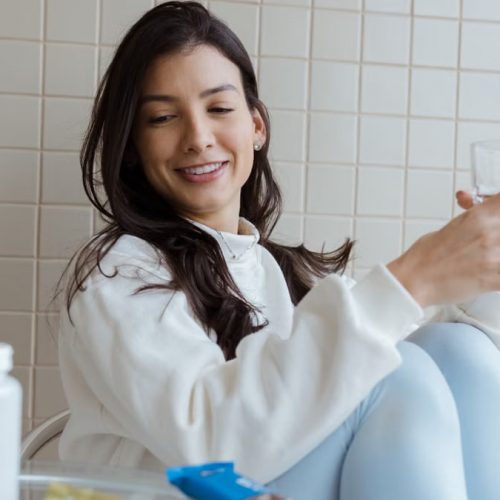 Smiling woman holding a glass while seated, with pill bottles on the table.