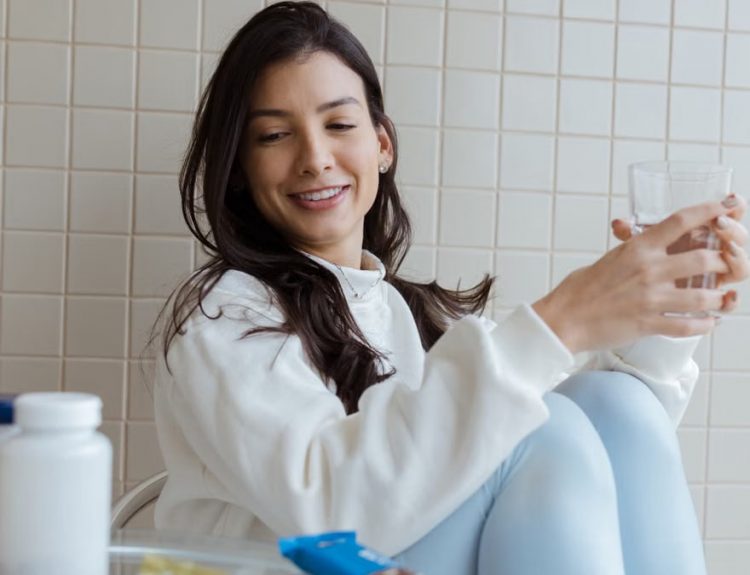 Smiling woman holding a glass while seated, with pill bottles on the table.